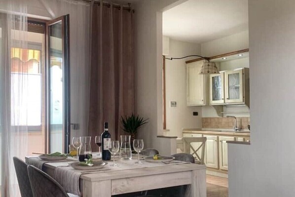 A dining area with a table set for four, a bottle of wine, and several glasses. A plant is on the table, and the room has a view of a kitchen with cabinets and a hanging light fixture.