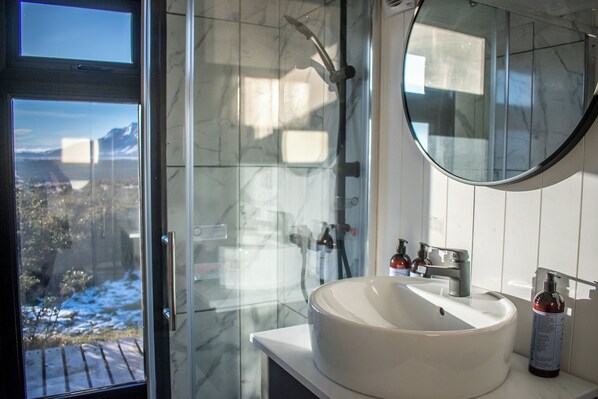 A bathroom with a large window, a white sink, and a shower with a glass door. - Tiny Homes Villa Cariño (Natales)