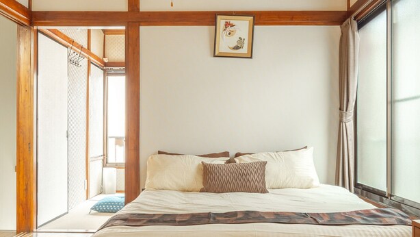 Quarto japonês tradicional com teto de madeira, uma cama grande com roupa de cama branca e uma porta deslizante que dá para uma varanda.