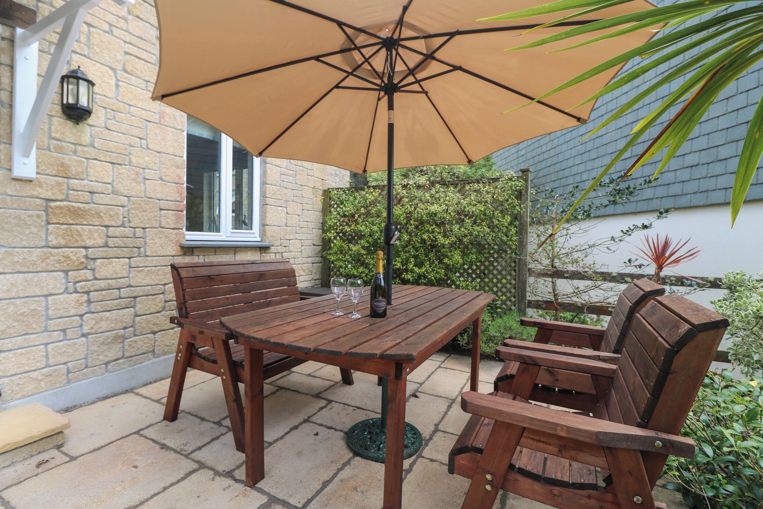 Une table de patio en bois, surmontée d’un parasol beige, deux chaises en bois et une bouteille de champagne.