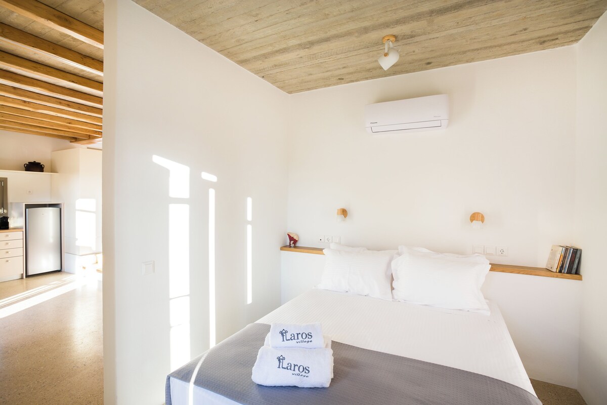 A hotel room with a bed, a bedside table, a shelf with books, an air conditioning unit, and a kitchen area visible through an open door.