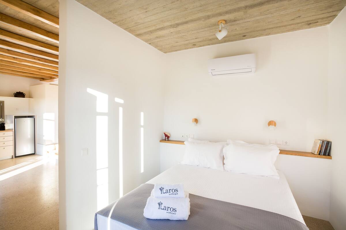 A hotel room with a bed, a bedside table, a shelf with books, an air conditioning unit, and a kitchen area visible through an open door.