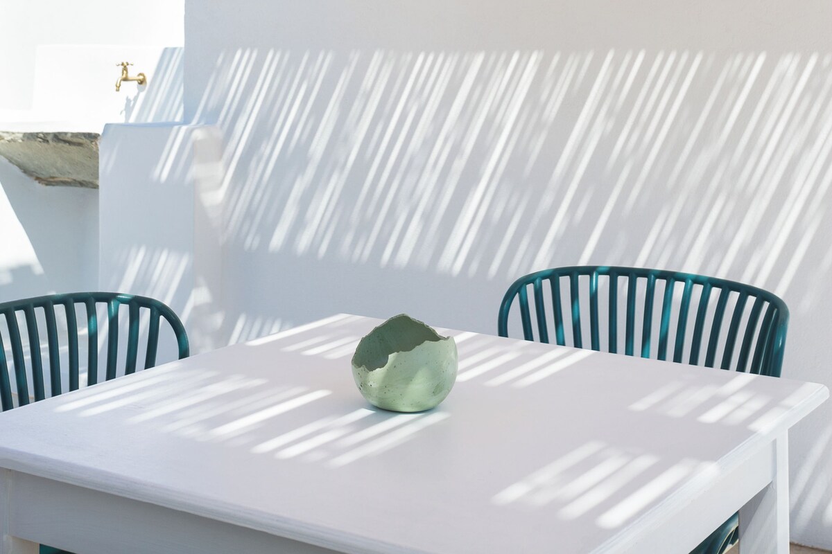A white table with a green bowl, two teal chairs, and a black pendant light under a wooden ceiling.