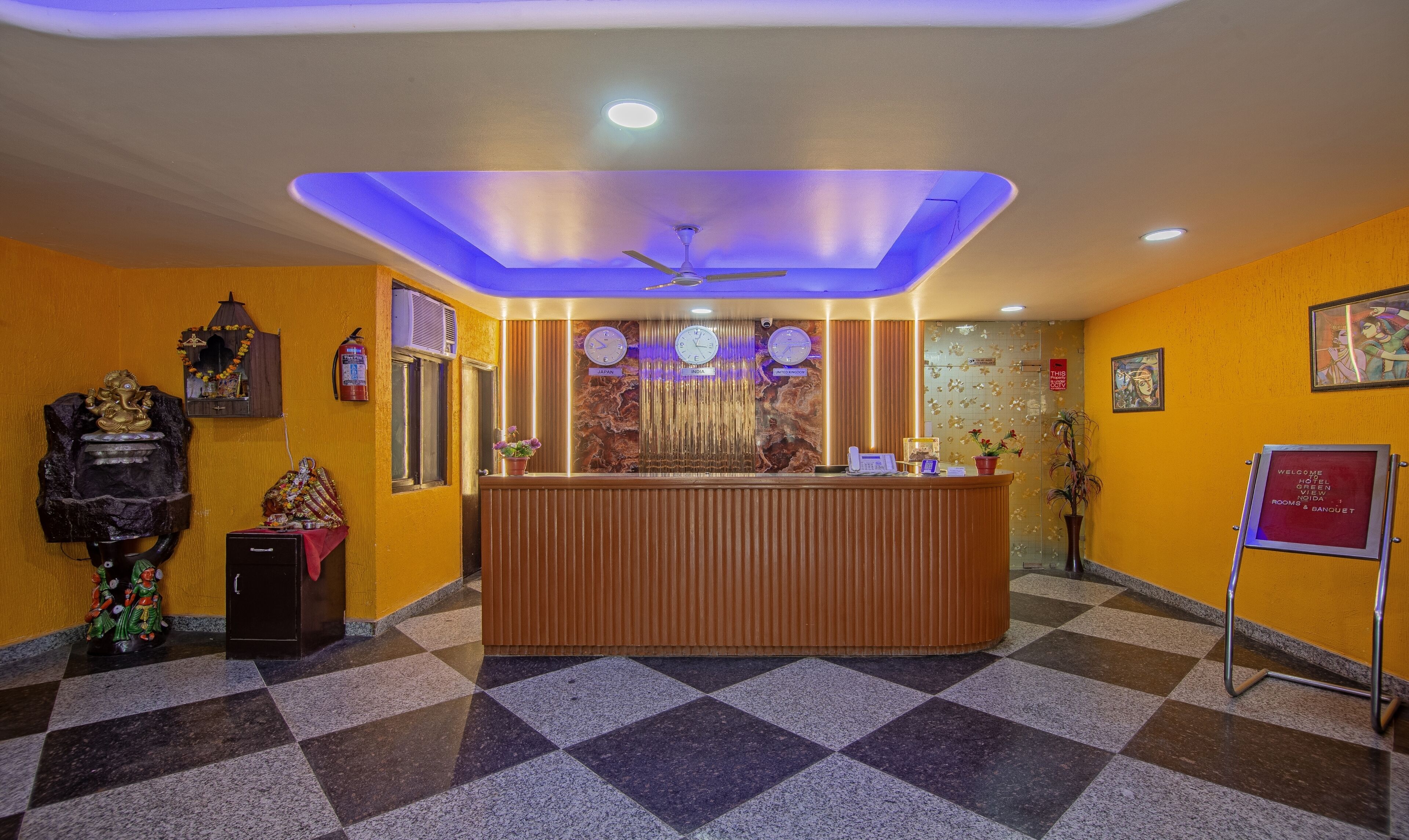 A hotel reception area with a check-in counter, a ceiling fan, and a clock displaying time for Japan, India, and the United Kingdom.