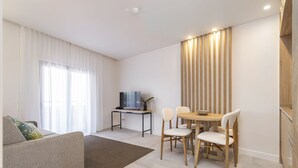 Una sala de estar moderna con un sofá, una mesa de comedor con sillas, un televisor sobre un mueble bajo y un ventanal con cortinas.
