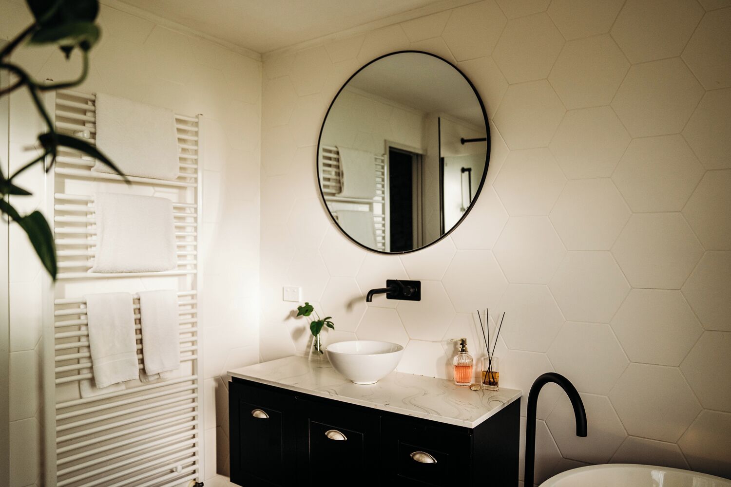A bathroom with a round mirror, white hexagonal tiles, a white sink, black cabinets, and a towel rack with towels.