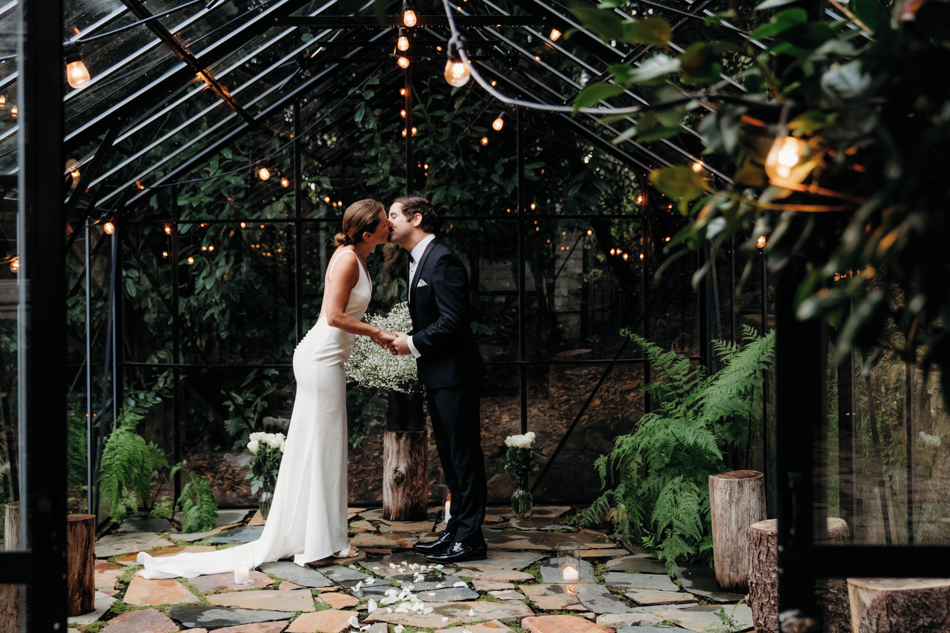 A couple kissing under a canopy with string lights, surrounded by greenery and stone flooring.