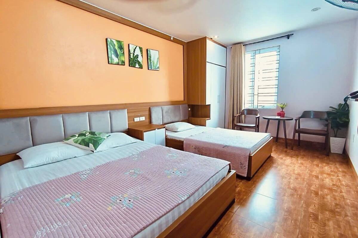 A hotel room with two beds, a small table with chairs, a window with curtains, and a ceiling fan.