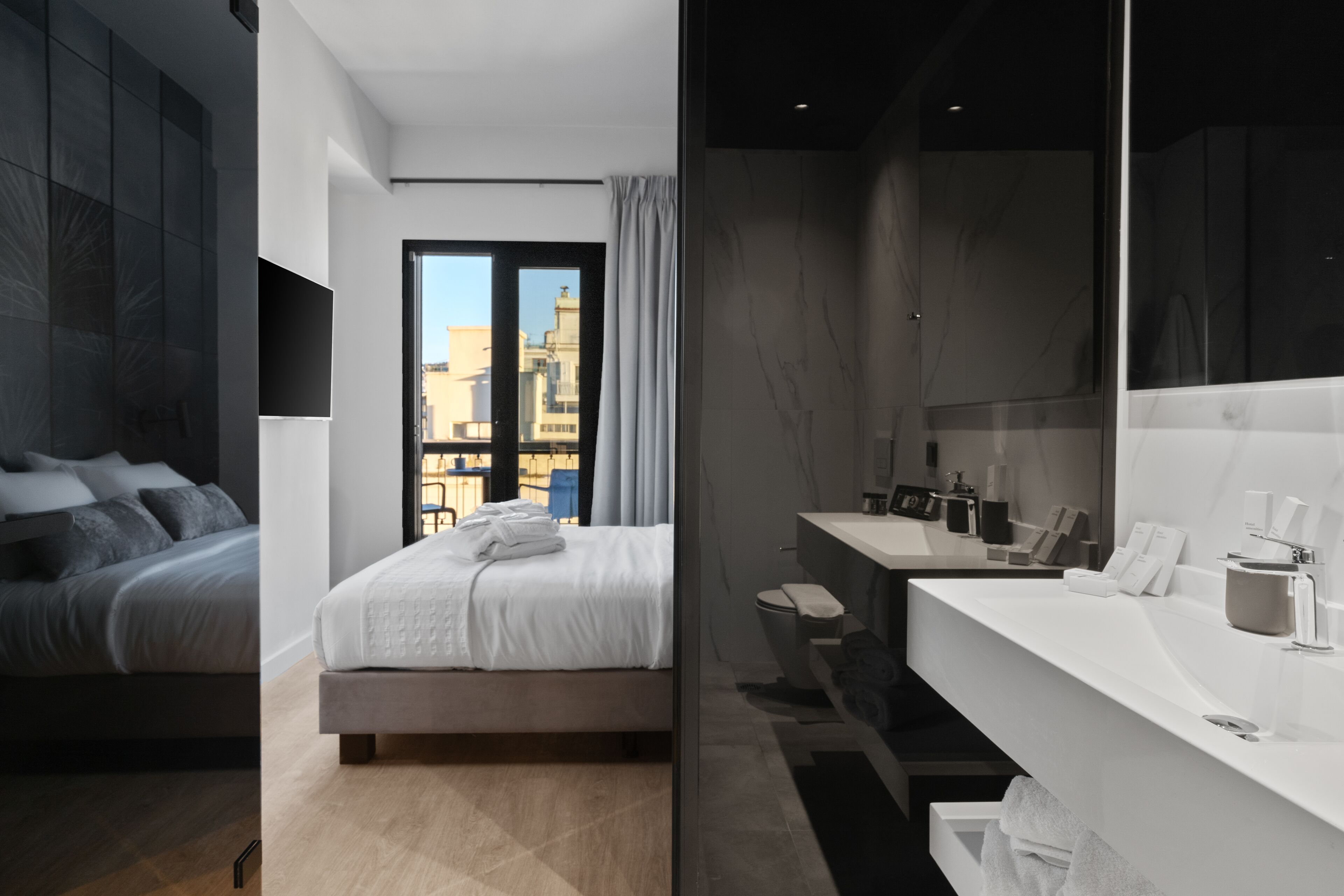 A modern hotel room with a large bed, a bathroom with a vanity, and a view of the city through a large window.
