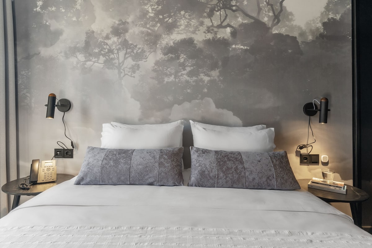 A neatly made bed with white linens, two grey pillows, and a wall mural of trees. There are two bedside tables with lamps and a telephone on one.