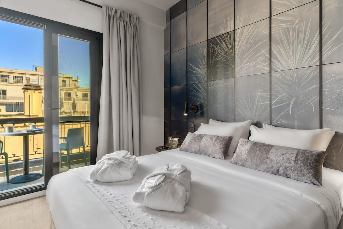 A modern hotel room with a large bed, two towels, a nightstand, and a view of the city through a large window.