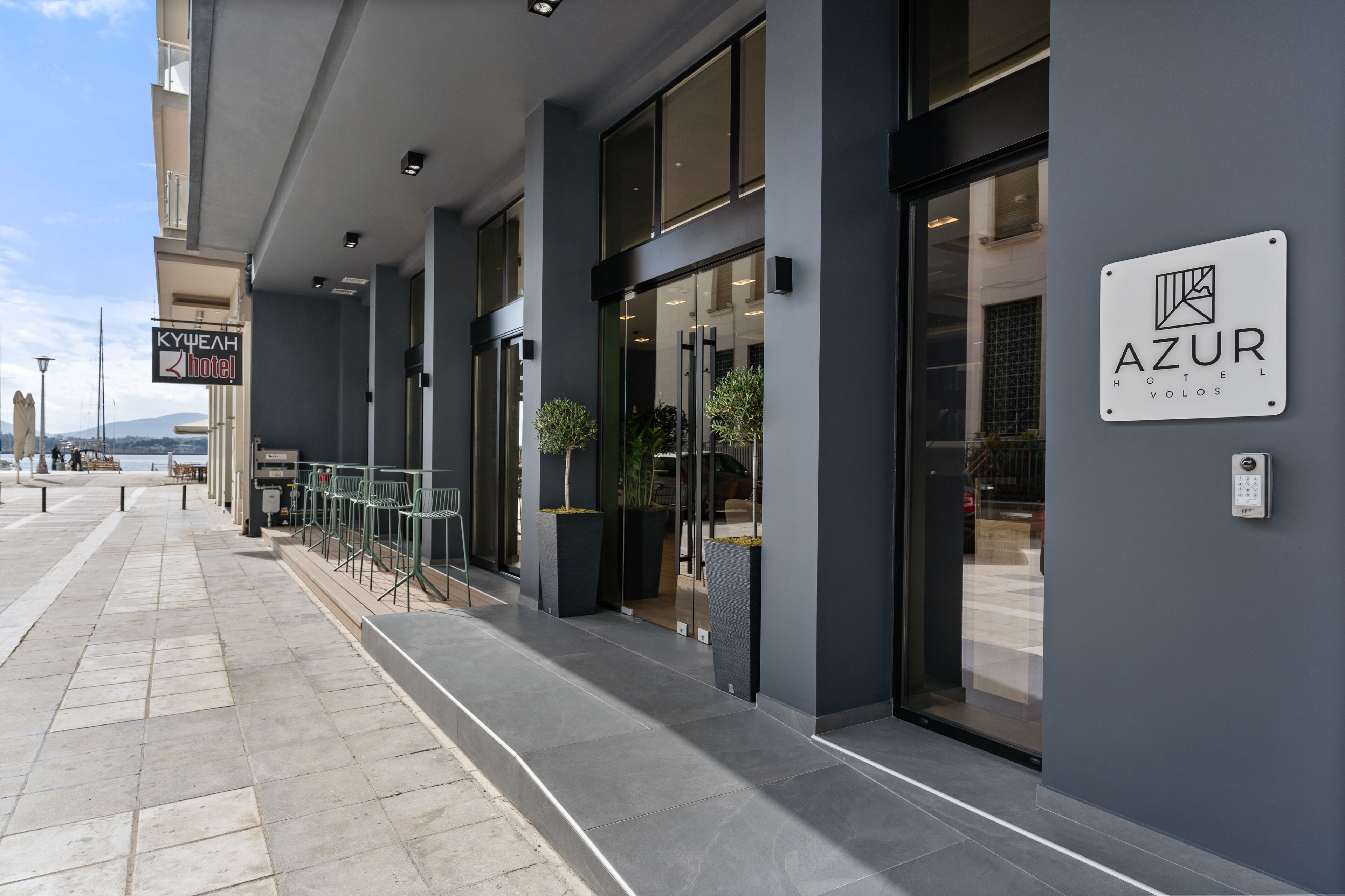 Una entrada de hotel moderna con puertas de cristal, asientos exteriores y un letrero que dice 'AZUR HOTEL VOLOS'.