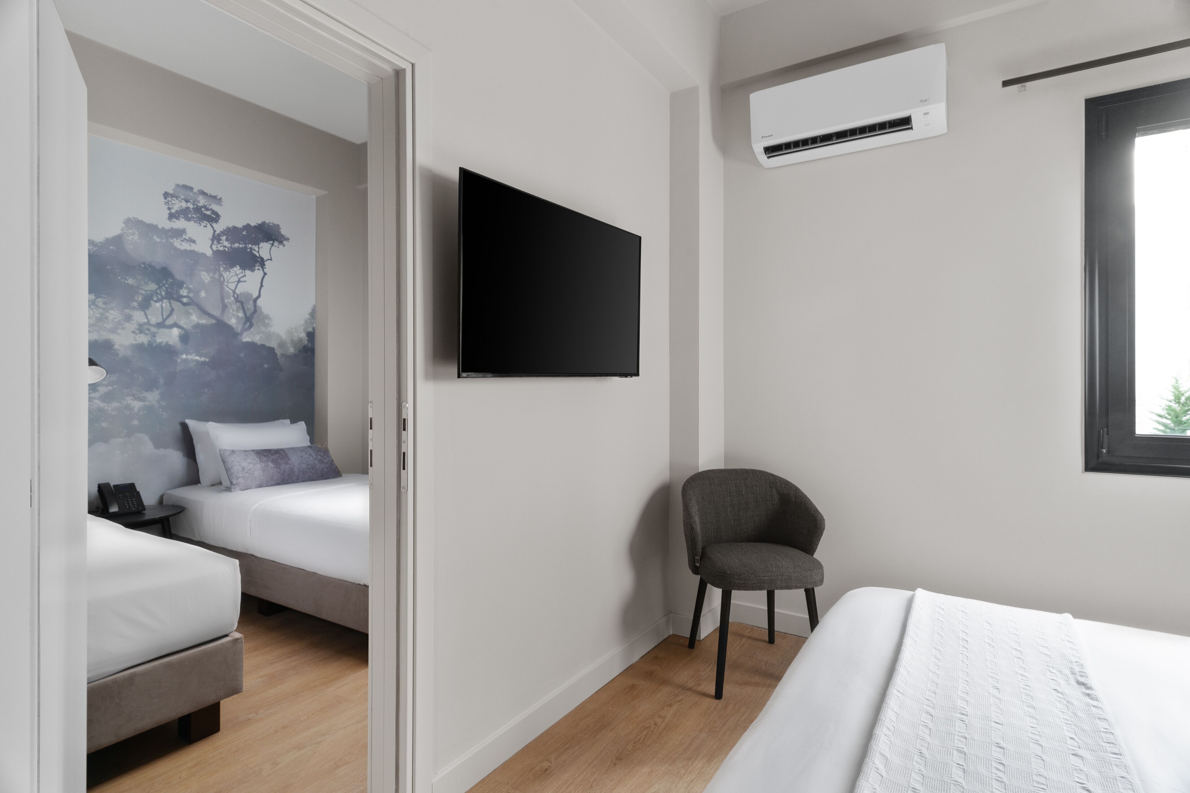 A modern hotel room with a bed, a chair, a flat-screen TV, and a wall-mounted air conditioner.