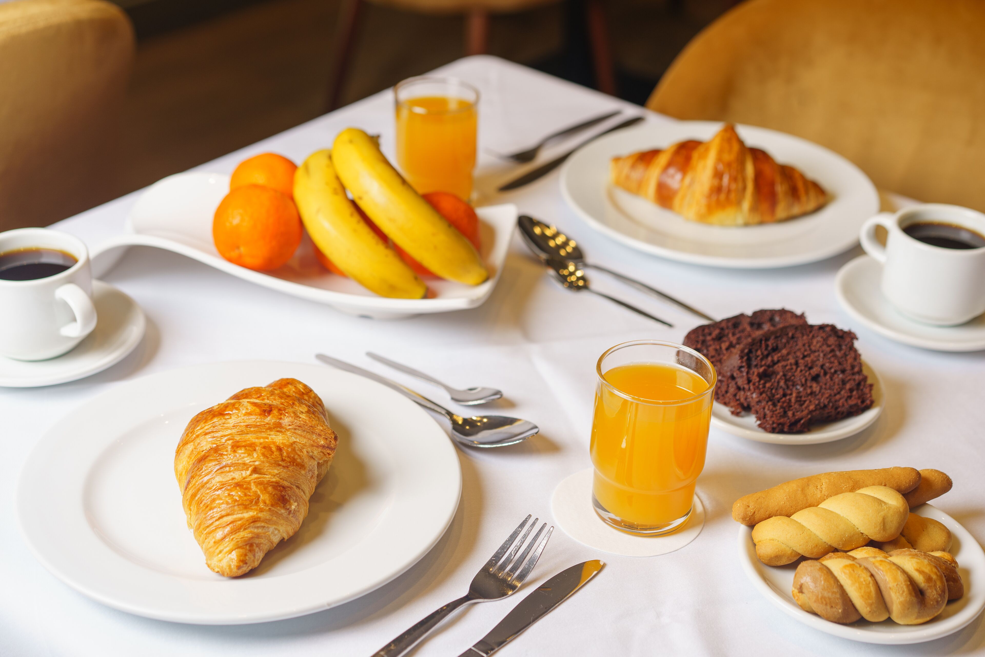 Desayuno con croissants, plátanos, naranjas y un vaso de zumo de naranja sobre una mesa blanca.