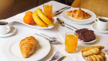 Desayuno con croissants, plátanos, naranjas y un vaso de zumo de naranja sobre una mesa blanca.