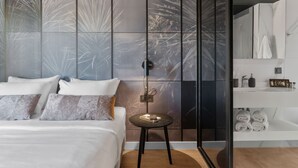 Een moderne slaapkamer met een groot bed, een klein tafeltje en een badkamer met spiegel en handdoekrek.