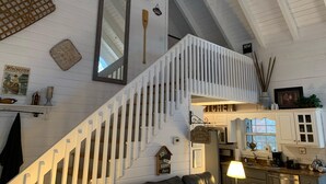 Un ambiente spazioso con soffitto alto, pareti di legno bianco e una scala con ringhiere bianche, una zona cucina e un'area pranzo con tavolo e sedie.