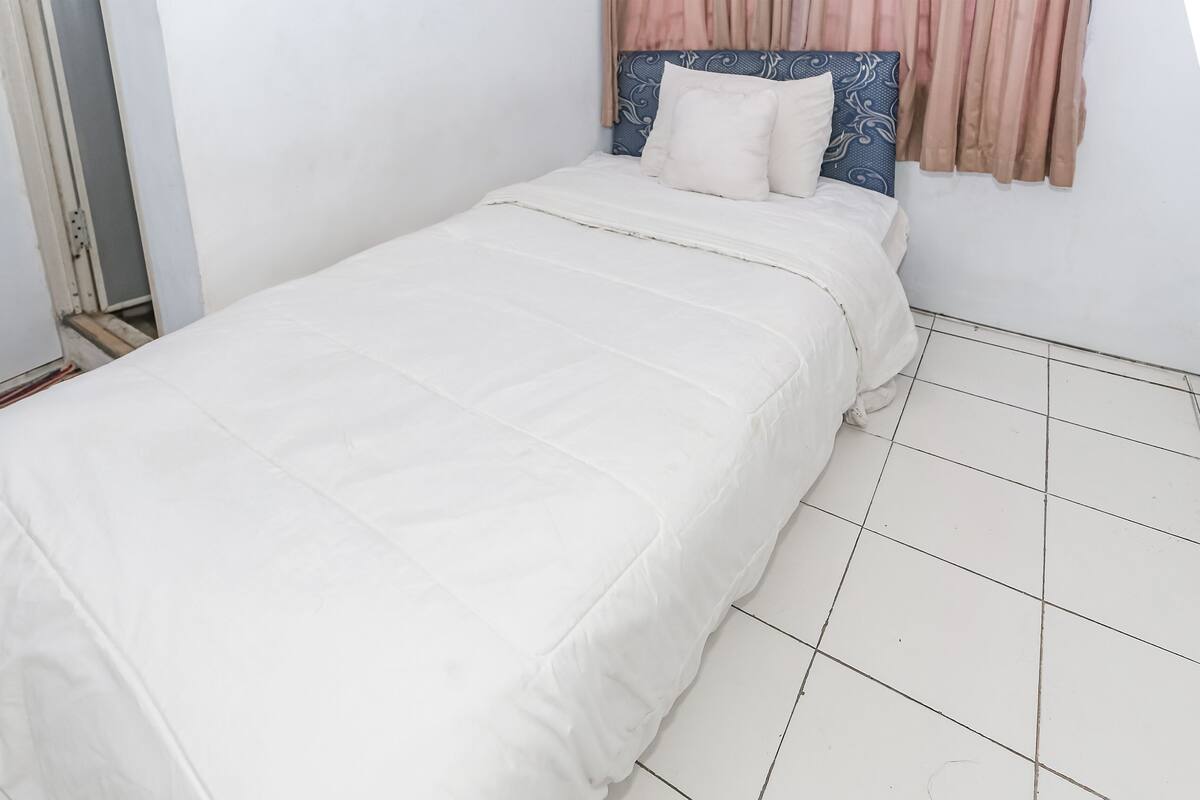 Un lit simple avec un matelas et des oreillers blancs, une tête de lit à motifs bleus et blancs, et un sol carrelé.