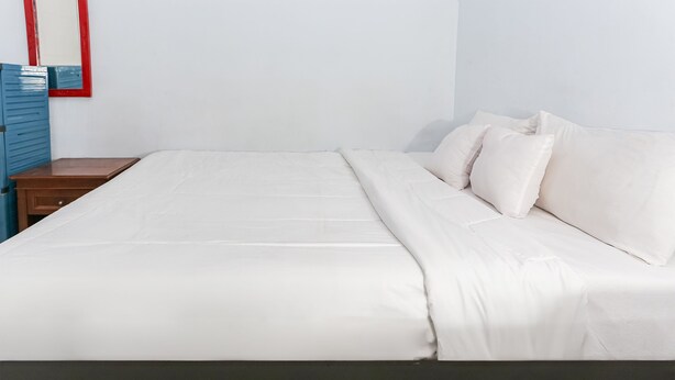 Uma cama bem arrumada com lençóis e almofadas brancos, uma mesinha de cabeceira de madeira com uma gaveta e um espelho com moldura vermelha na parede.