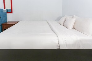 Een net opgemaakt bed met witte lakens en kussens, een nachtkastje van hout met een lade, en een spiegel met rode lijst aan de muur.