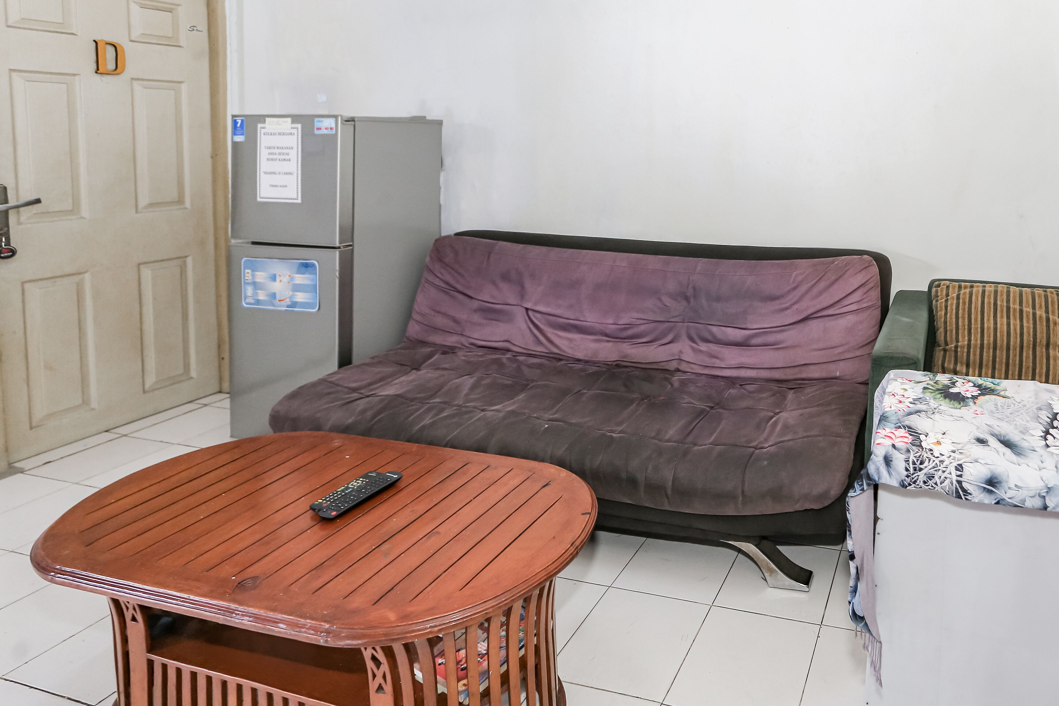 A small room with a wooden table, a sofa, a refrigerator, and a door marked with a letter.