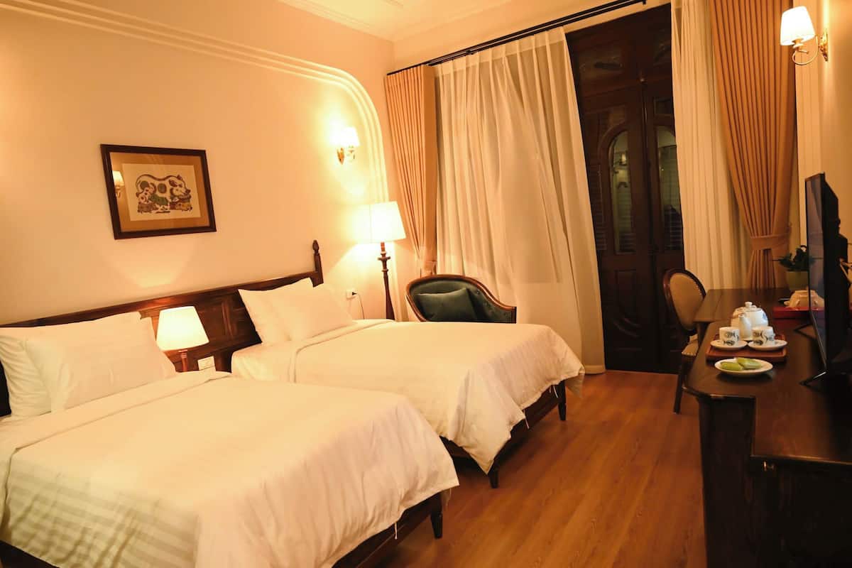Une chambre d’hôtel avec deux lits, un bureau avec une lampe, une chaise et une table à manger mise avec des tasses à thé.