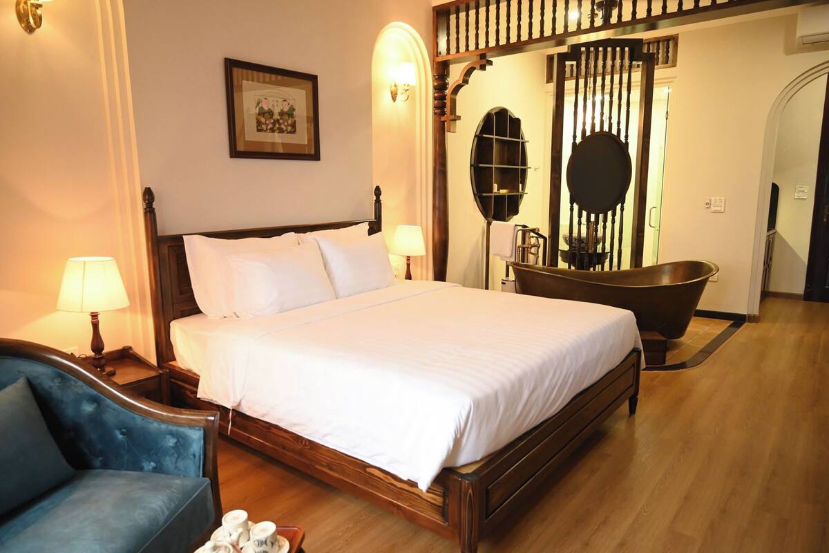 Une chambre d’hôtel avec un grand lit, un fauteuil bleu, une baignoire et un miroir fixé au mur.