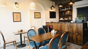 Interior de un restaurante con una mesa de comedor de madera, sillas tapizadas en azul, lámparas colgantes y una zona de bar con estantes llenos de botellas y vasos.