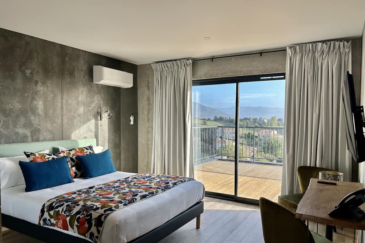 A modern hotel room with a large bed, a balcony with a view, and a small table with a chair.