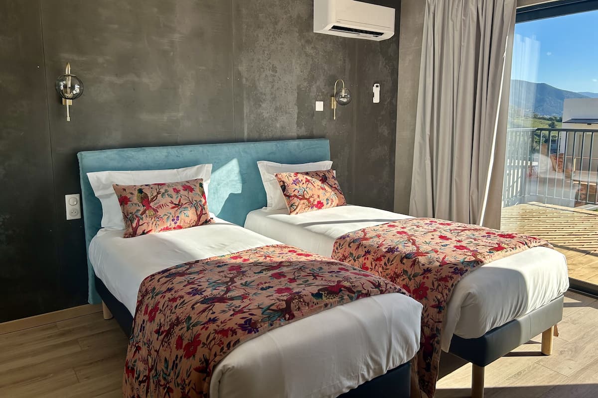 A hotel room with a large bed, two bedside tables, a window with a view, an air conditioning unit, and a curtain.
