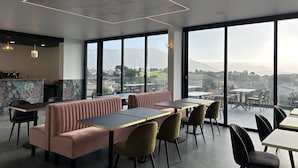 A modern restaurant with a high ceiling, large windows offering a view of the city and mountains, and a bar area with a floral wallpaper design. - Hôtel Casa Matteu (Afa)
