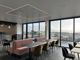 Un restaurante moderno con techos altos, amplios ventanales que ofrecen vistas a la ciudad y a las montañas, y una zona de bar con un diseño de papel tapiz floral.