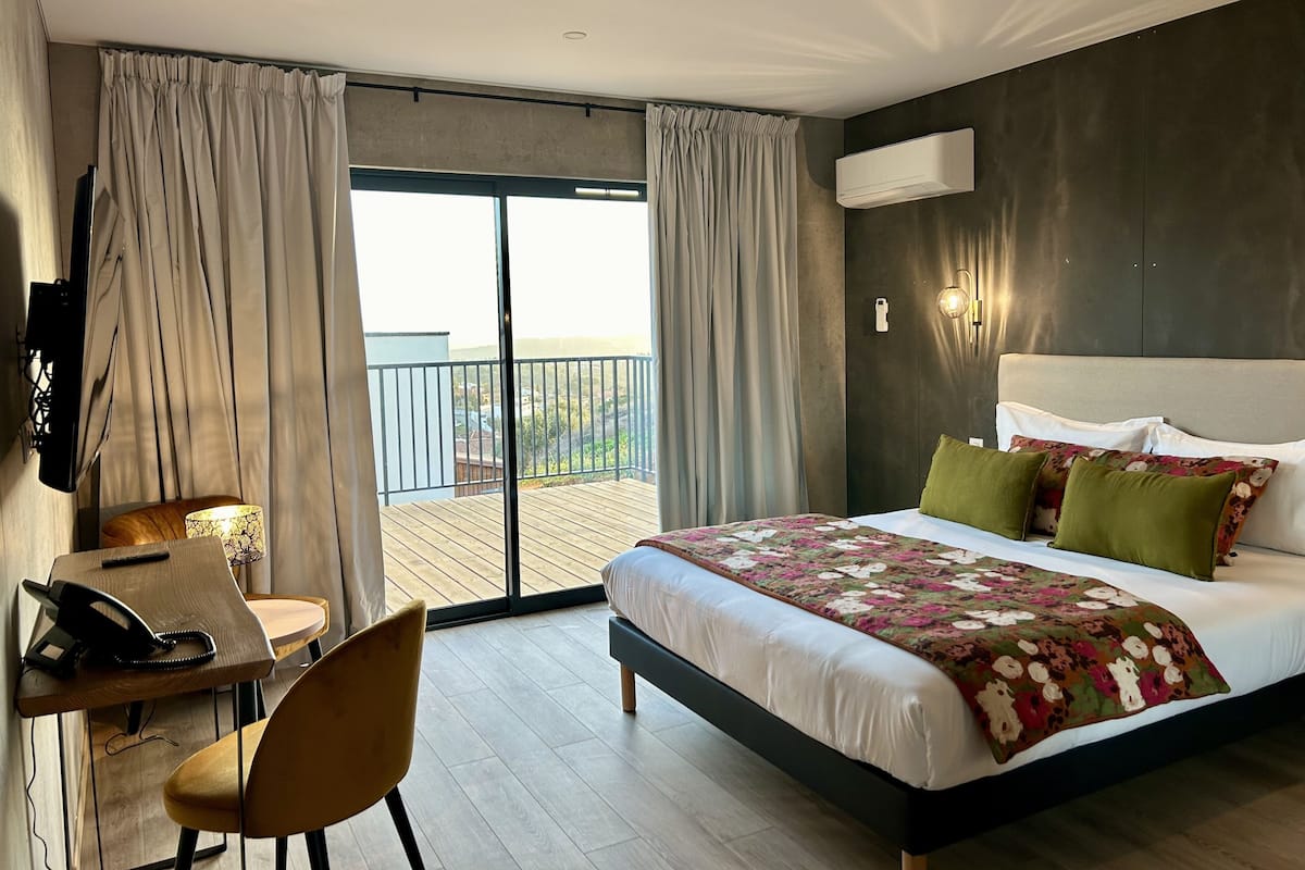 A hotel room with a bed, a desk, a chair, a balcony with curtains, and a view of the outside.