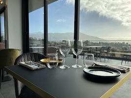 Mesa dispuesta con platos, vasos y cubiertos, con vistas a un paisaje con montañas y una masa de agua.