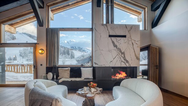 Una habitación acogedora con chimenea, ventanas amplias con vistas a una montaña nevada y un espacio para sentarse confortable.