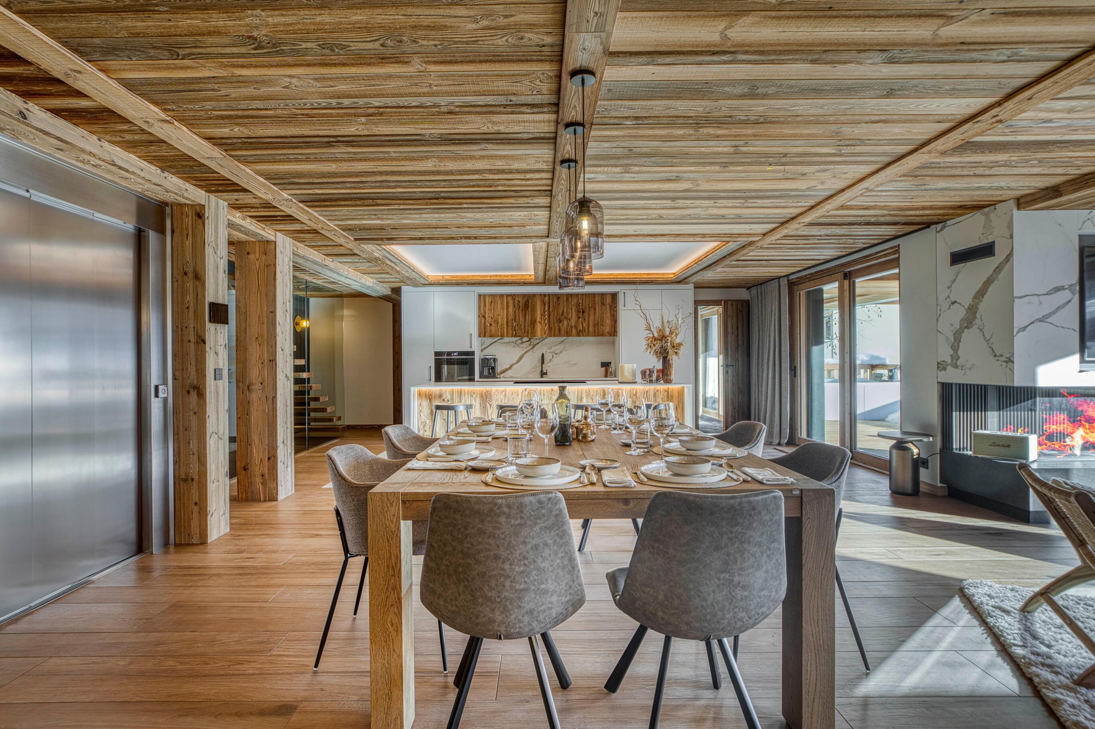 A modern dining area with a wooden ceiling, a long table set for six, and a kitchen in the background.