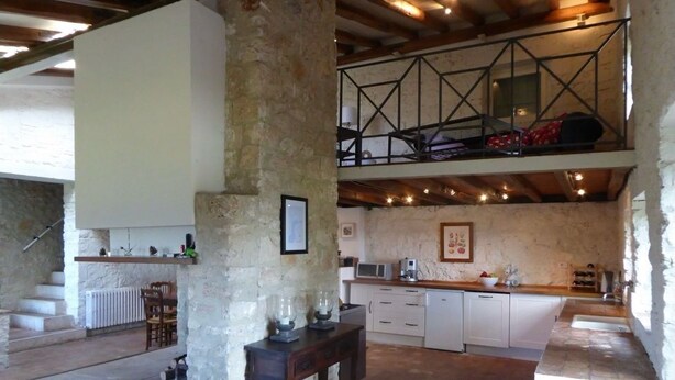 Un interior rústico con pared de piedra, vigas de madera, una cocina con armarios blancos y una zona de comedor con mesa y sillas.