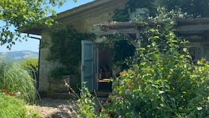 Una casa de piedra rústica con chimenea, un jardín con diversas plantas y flores, y un cielo azul despejado.