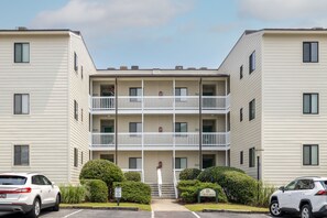 A multi-story apartment building with a central courtyard, balconies on each floor, and a parking lot with two cars. - Point Emerald B-305, Almost Oceanfront Condo in Emerald Isle, NC! (Emerald Isle)