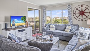 Een woonkamer met een televisie, bank, salontafel en een groot raam met uitzicht op huizen en groen.