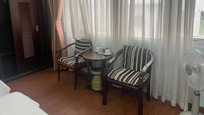 Una habitación de hotel con una cama, dos sillones a rayas, una mesita redonda pequeña con una taza y un plato, un ventilador y cortinas traslúcidas.
