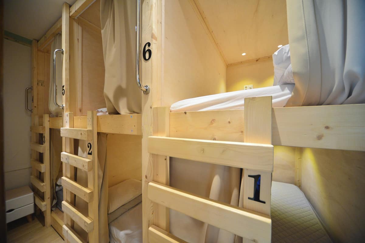 Reihenweise hölzerne Etagenbetten mit den Nummern 1 bis 6, jeweils mit einer Klapptreppe und einer weißen Matratze.