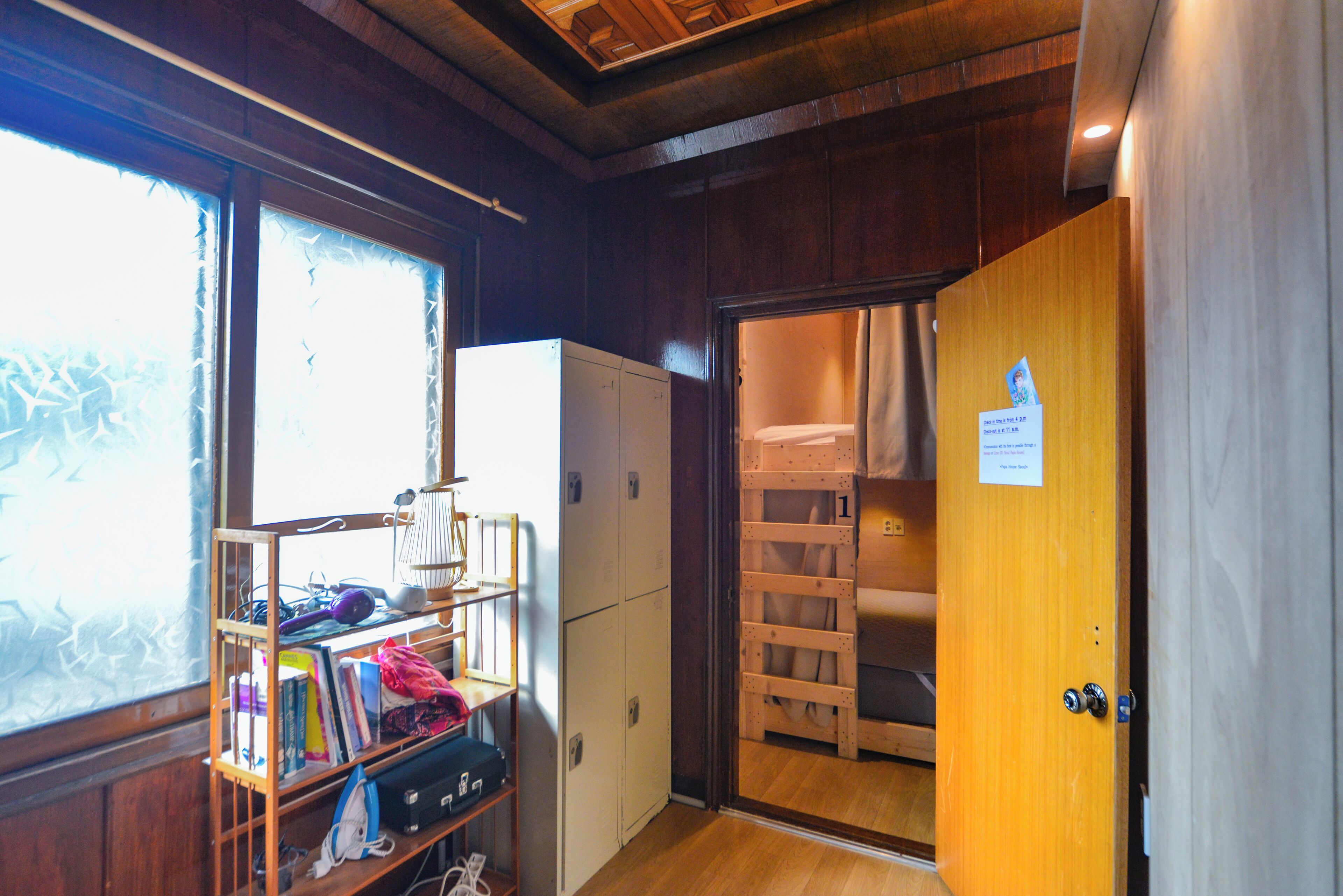 A room with a wooden ceiling, a window with frosted glass, a white refrigerator, a wooden door with a sign, and a shelf with books and a lamp.