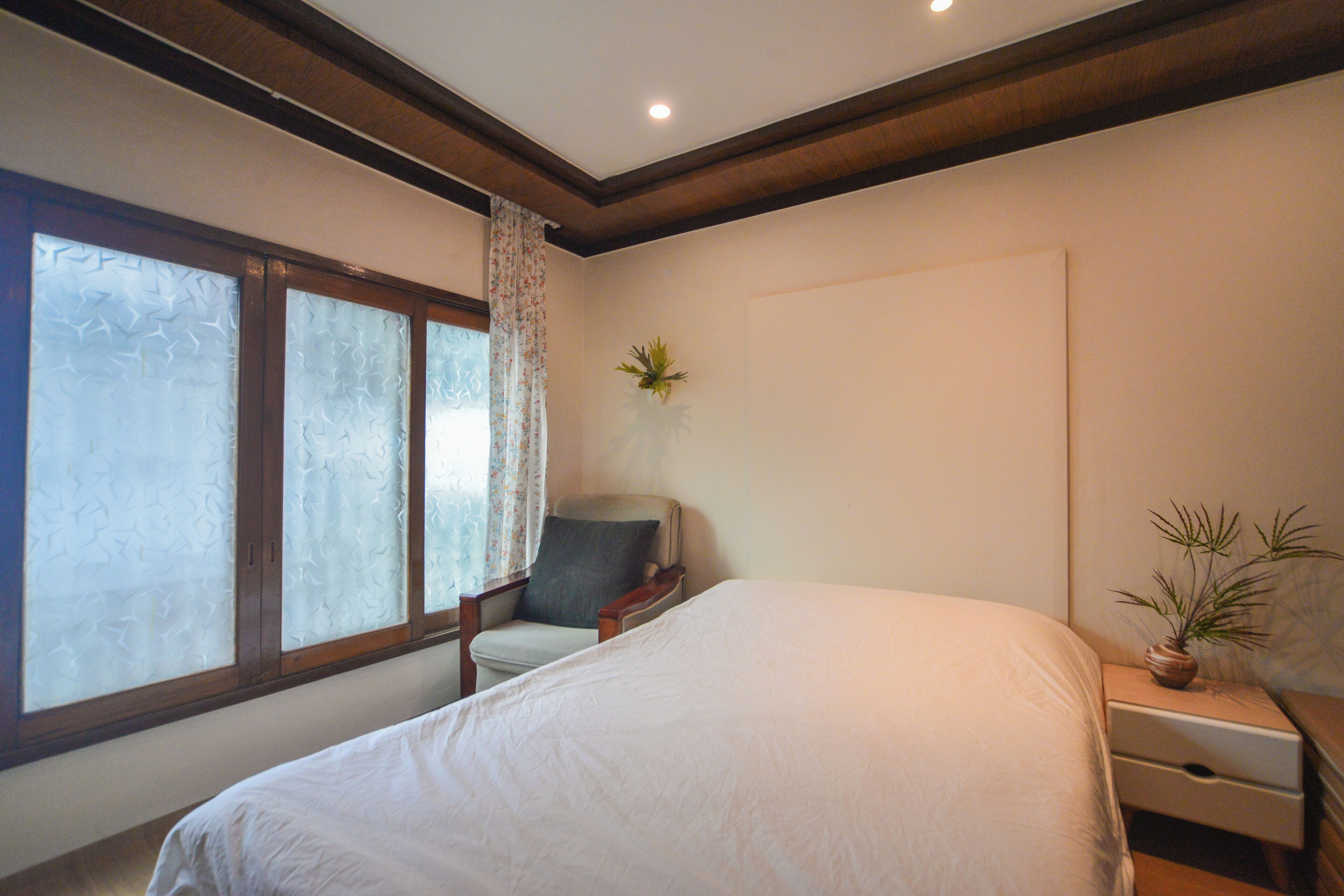 A bedroom with a large bed, a chair, a nightstand with a plant, and a window with frosted glass.