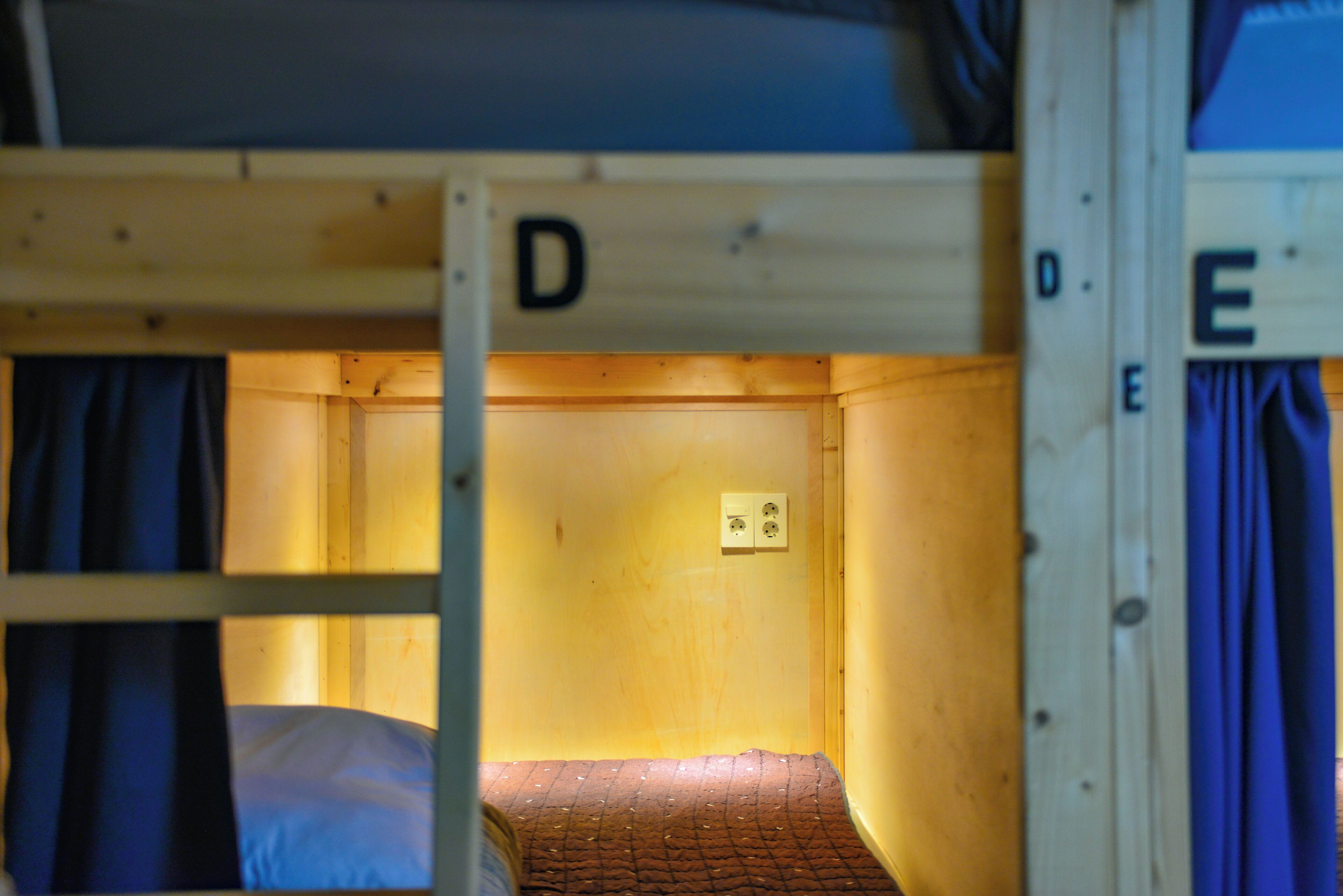 Habitación con literas de madera, una de ellas identificada con la letra 'D', y una cama individual con colchón y manta.