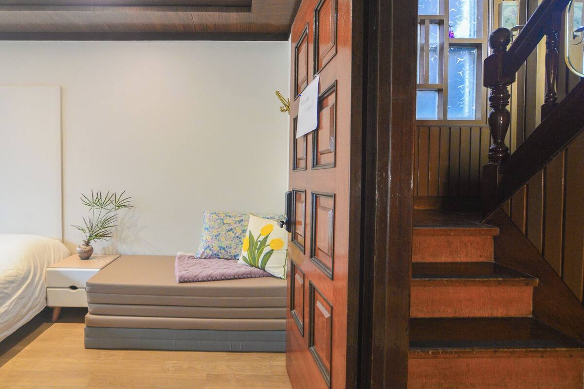 Ein Schlafzimmer mit einem Bett, einem kleinen Nachttisch, einer Topfpflanze und einem Fenster mit Blick ins Freie.