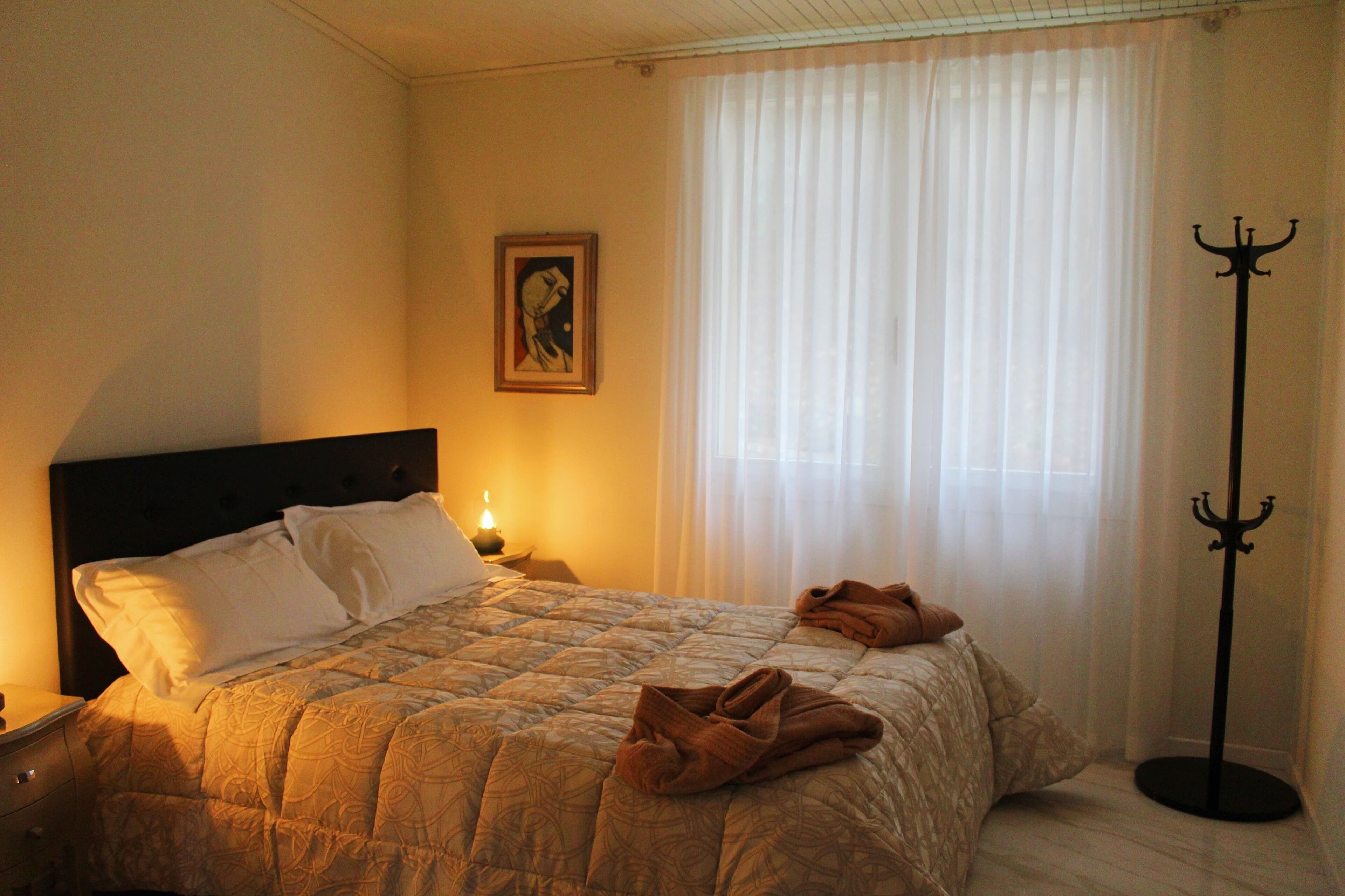 A neatly made bed with a quilt, pillows, and folded towels. There is a nightstand with a lamp and a framed picture on the wall. A coat stand is visible in the corner.