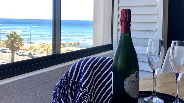 Eine Flasche Miss O'Connell's Sekt auf einem Fensterbrett mit zwei Gläsern, Blick auf einen Strand mit Palmen und Autos.