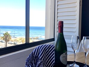 A bottle of Miss O'Connell's bubbly on a windowsill with two glasses, overlooking a beach with palm trees and cars. - Studio Blue - Mouille Point, from beach to bar! With sea and yachts in the bay.
 (Cape Town)