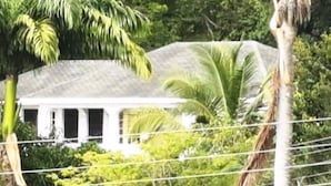 Un bâtiment blanc surmonté d’un toit gris, entouré d’une végétation luxuriante et de palmiers.